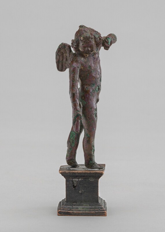 A winged child stands with one elbow raised in this cast bronze, free-standing sculpture. The surface is rough, pitted, and oxidized to green in many areas. In this photograph, the boy’s body faces our right in profile. His head tips down over his left shoulder, coming toward us, and his far elbow is raised as if to shield his face. His right arm hangs long by his side. He has short curly hair and his features are smoothed. A knubby wing thrusts back from the shoulder closer to us. With rounded feet, the child stands on a pedestal base rubbed gold across moldings along the top and bottom.