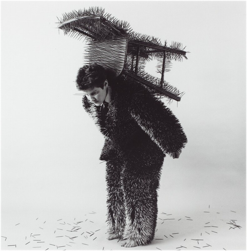 A person stands and bows forward with a chair on their back in this vertical, black-and-white photograph. The person’s clothing and chair bristle with a carpet of needle-like spikes. The person looks down, their body angled to our left as the arms stretch back by their sides. They have pale skin and short, dark hair. They wear a suit and tie and are barefoot. The chair is balanced on the person’s back with the seat facing forward at the back of their neck, so the back of the chair seems to open like a gaping mouth. More spikes are scattered on the white floor in front of a white background.