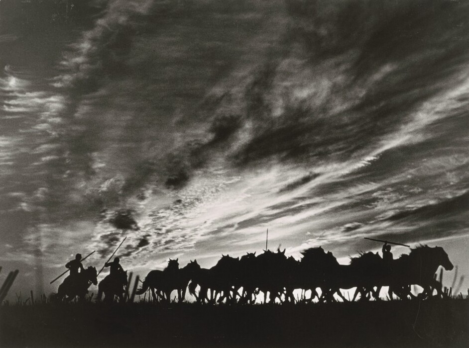 The image shows silhouettes of horse riders passing along the low horizon. The dark figures on horseback are visible against swirling clouds in the vast sky, with a monochrome color palette of deep blacks and various shades of gray. Lighting suggests a time close to dusk or dawn, enhancing the visual impact.