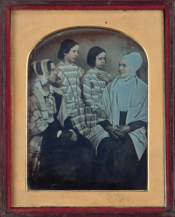 The image depicts four individuals, from the waist up. An elderly woman is seated with her hands clasped, while three young women stand behind her. The elderly woman wears a light bonnet and shawl. The young women have dark hair styled to reveal their ears, wearing similar plaid dresses with high collars. The women have different facial expressions. The photograph is in an ornate wooden frame with a decorative matte. The background is plain, focusing attention on the individuals.