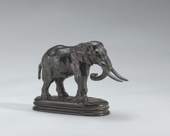 The sculpture features an elephant standing on a small, rounded rectangular base. It is made of dark material, likely bronze, with a polished finish. The elephant has detailed features such as tusks, ears, eyes, and trunk. The sculpture is small-scale relative to a real elephant and is suitable for decoration or art display. The dark color and smooth surface accentuate the elephant's form and details.