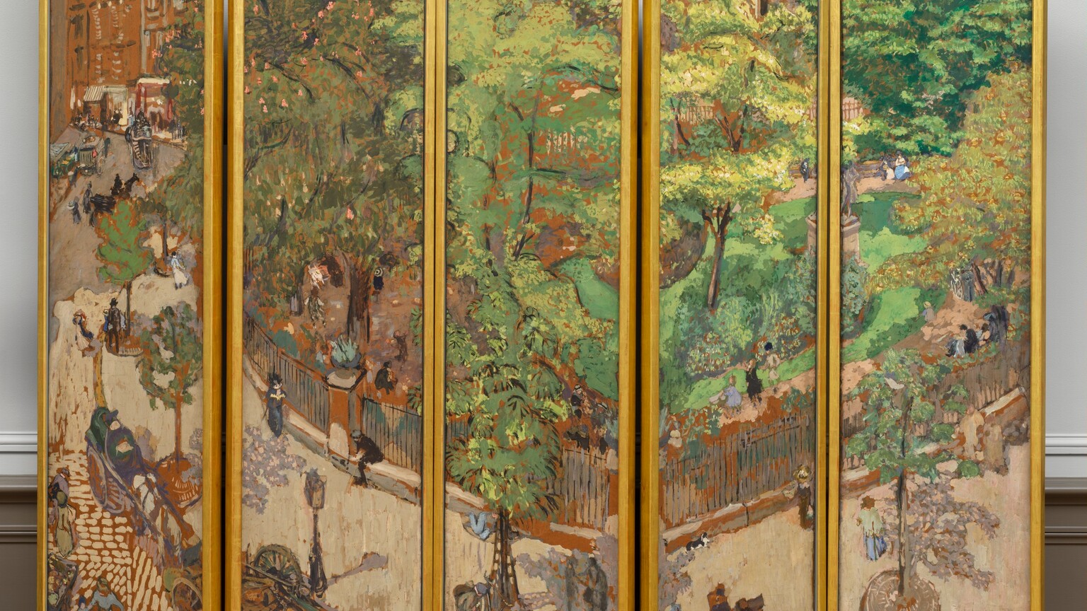 A screen made up of five tall, rectangular panels, set side by side and each surrounded by a gold frame, is painted as a single scene showing a tree-lined sidewalk curving around a park in a city. The scene is loosely painted with short, rounded brushstrokes. The top two-thirds to three-quarters of most of the panels are filled with the lime and olive-green leaves of the trees that line the sidewalk and park. In the leftmost panel, the sidewalk and road lead back to a row of caramel-brown building façades. The sidewalk is pale taupe, and the street is painted with dashes of the same taupe against terracotta brown, suggesting cobblestones. Spindly trees are spaced in a row along the sidewalk in round holes covered with smoke-gray metal grates. A black fence, painted with thin, sometimes broken black lines encloses the park beyond, which has a path around plantings and the vivid green lawn. Touches of pink on a sage-green tree to our left in the park suggest flowers. A gray statue on a high plinth is partially lost in the break between the two rightmost panels. Men, women, and children, painted with a few strokes of black, gray, or marine or periwinkle blue, walk along the sidewalk and the garden path, or sit at the base of the fence or on benches spaced along the sidewalk. The women seem to wear long dresses and the men dark clothing and hats. Two carriages are pulled up on the street near a lamp post alongside the sidewalk near the lower left. In the leftmost panel, horse-drawn carriages move along the road leading back to the buildings, and more people seem to be gathered on the sidewalk near the left edge of the panel in the distance. The artist signed the work with brown paint in the lower right corner: “E. Vuillard.” The panels of the screen have been set up so the panels rest on a platform or on the floor in a shallow zig-zag pattern, in a room with an off white wall and bisque-brown molding along the floor.