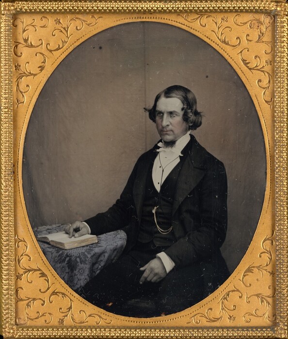 The image features a man seated from the waist up. He is slightly turned to the side, facing forward with his gaze towards the camera. The man has distinct facial features, clean-shaven with a small patch of facial hair below his lower lip. His hair is styled with a middle part and combed back on the sides. He is dressed in a dark suit with a waistcoat and white shirt, along with a bowtie. A chain attached to his waistcoat indicates a pocket watch. Leaning on a table with a patterned cloth, he has an open book in front of him. The background is plain, and the photograph is framed in gold with floral patterns.