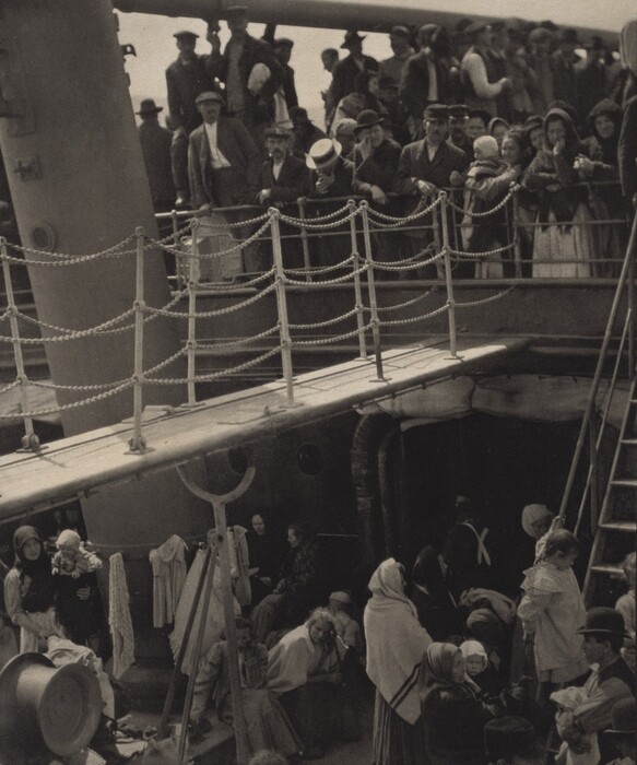 The image shows individuals on a ship's deck. The deck is divided by a railing with ropes, and the upper levels show people leaning and observing. The ship's architecture, with a large structure cutting diagonally across the image and a ladder on the right side, suggests a strong maritime vessel. Various clothing styles are visible, indicating a diverse group ranging from well-dressed to simpler attire. The lighting is soft, creating a subdued yet distinct atmosphere, and the composition feels bustling with people engaged in various activities or conversations.