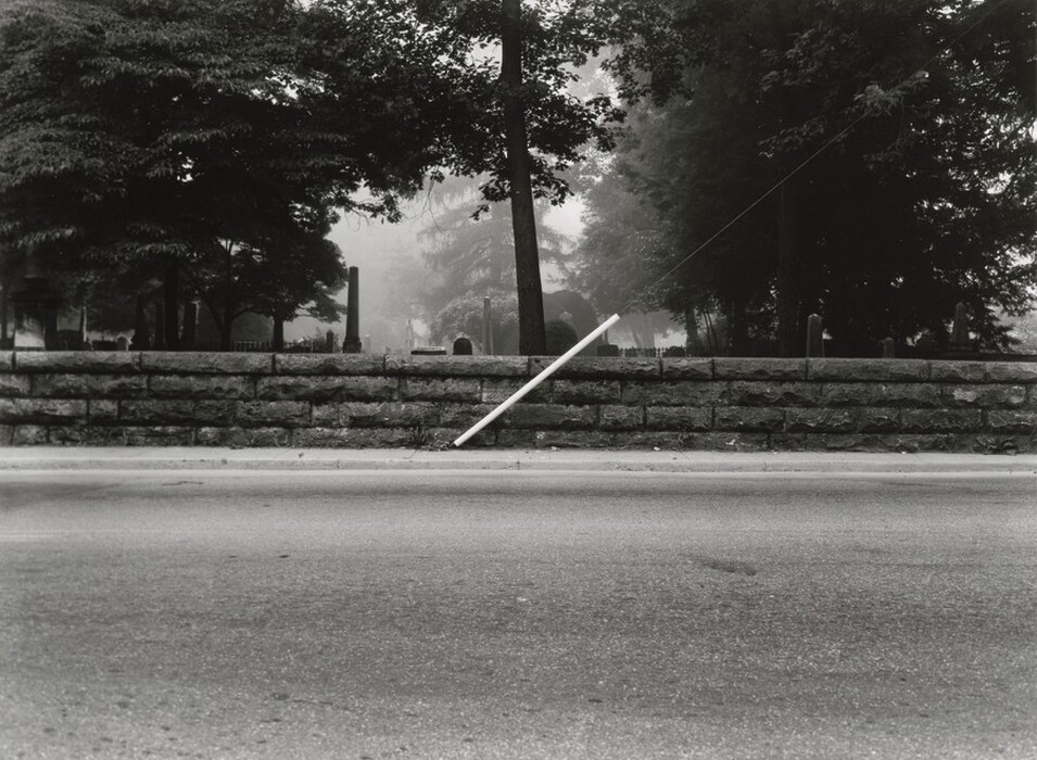 The image shows a street scene from a direct, eye-level point of view. An asphalt road leads to a stone wall, beyond which are tombstones and trees in a light fog. The horizon is marked by the wall, with grave markers and tree trunks rising against it. A thin pole leans diagonally into the scene. The image is in black and white, with a grayscale palette highlighting the contrast between the trees' dark shadows and the misty background.