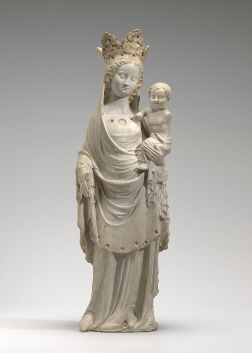 A crowned woman and the baby she holds on one hip are carved from white marble in this freestanding sculpture. The woman has a round face with a long nose and slightly smiling lips. Her long curly hair is parted down the middle and held back under a high crown made of spade-shaped acanthus leaves. A cloth covers her head and drapes over her shoulders and across her hips. Divots are carved along the lower hem of the cloth. The dress she wears underneath has three circular openings along the squared neckline, and pointy-toed shoes peek out from under the hem of the long skirt. She holds the stem of a now-broken frond in her right hand, to our left, by that hip. Her other hand supports the baby. He has a round face, short, wavy hair, wide-set eyes, chubby cheeks, a delicate nose, and his lips are parted. His torso is nude, and a cloth wraps around his hips and falls in folds around his bare feet. His left arm, to our right, is missing beyond the shoulder. He leans the other elbow against the woman’s chest, and the lower arm is missing beyond that. The sculpture is photographed against a pale gray background.