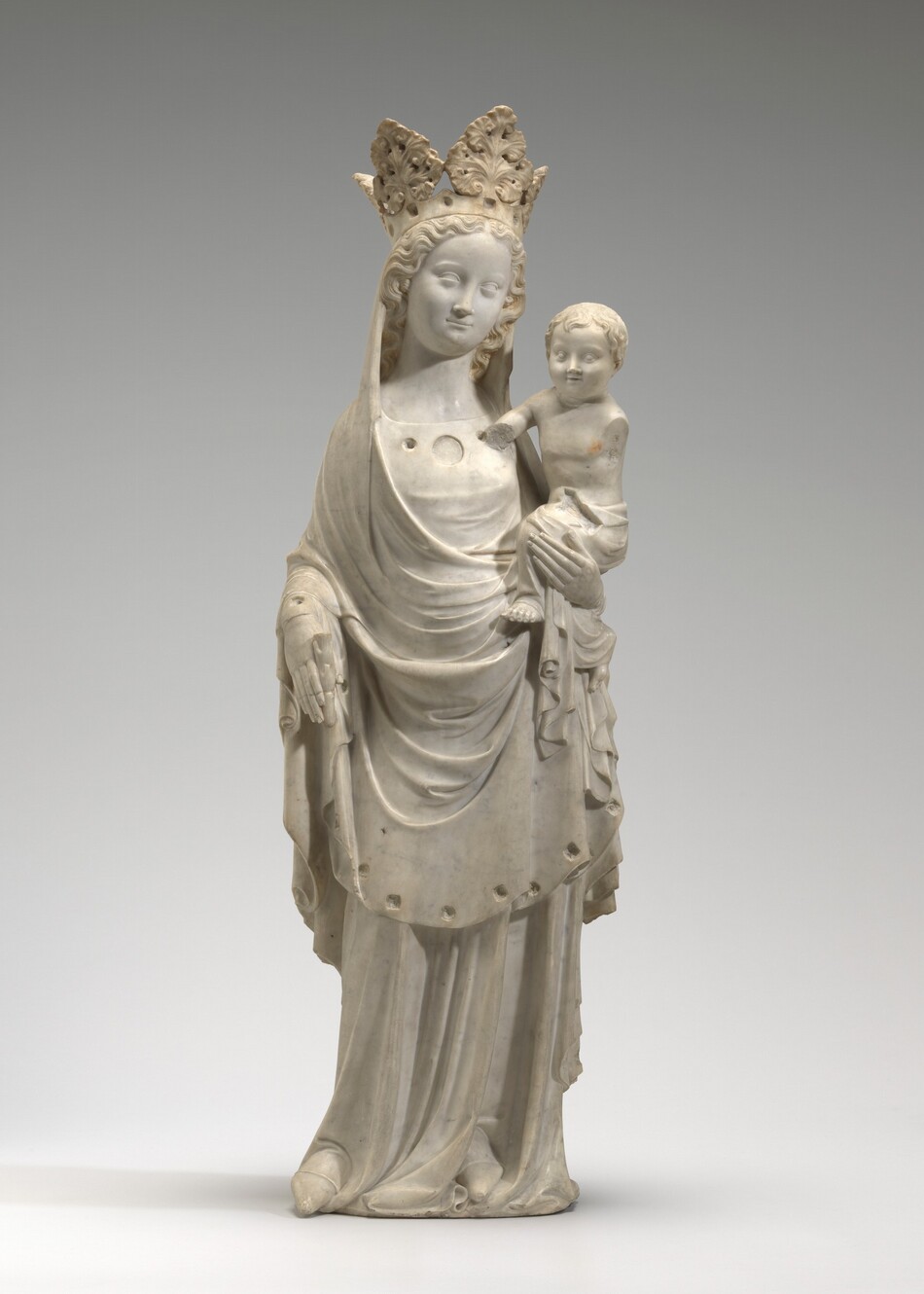 A crowned woman and the baby she holds on one hip are carved from white marble in this freestanding sculpture. The woman has a round face with a long nose and slightly smiling lips. Her long curly hair is parted down the middle and held back under a high crown made of spade-shaped acanthus leaves. A cloth covers her head and drapes over her shoulders and across her hips. Divots are carved along the lower hem of the cloth. The dress she wears underneath has three circular openings along the squared neckline, and pointy-toed shoes peek out from under the hem of the long skirt. She holds the stem of a now-broken frond in her right hand, to our left, by that hip. Her other hand supports the baby. He has a round face, short, wavy hair, wide-set eyes, chubby cheeks, a delicate nose, and his lips are parted. His torso is nude, and a cloth wraps around his hips and falls in folds around his bare feet. His left arm, to our right, is missing beyond the shoulder. He leans the other elbow against the woman’s chest, and the lower arm is missing beyond that. The sculpture is photographed against a pale gray background.