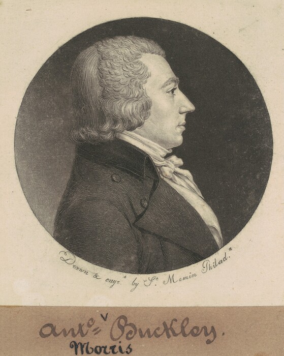 This image shows a detailed profile view of a man's upper body. He is positioned facing to the right, displaying a classical side portrait pose. His facial features include a prominent nose and a fully visible chin, with tightly curled hair that appears to be neatly styled, reaching down to the nape of his neck. He is dressed in formal attire, wearing a dark coat with visible buttons and a high white cravat wrapped around his neck. The background consists of a plain circular backdrop. Below the circular portrait, there is a section with the artist's signature, "Drawn & engd. by St. Memin Philad.," and some handwritten text on the lower portion.