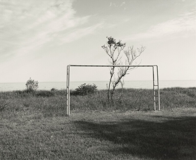 An angular jungle gym sticks out of a grassy lawn like an oversized staple in this horizontal black and white photograph. We are off to the left side so the horizonal bars on each narrow end are visible. On the far side of the jungle gym, a thin V-shaped tree with a puff of a canopy on one side grows up and over the equipment. The horizon of a pale body of water comes halfway up this photograph, and a few thin clouds sweep across the sky.
