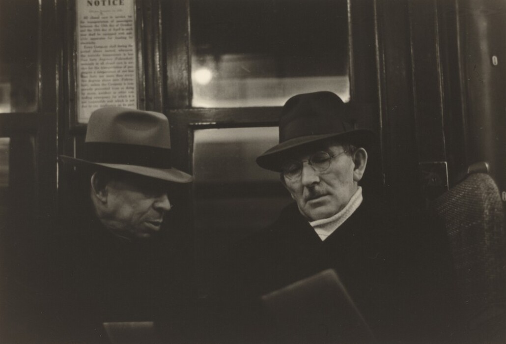 The image depicts two men sitting side by side. The man on the left is turned slightly towards the other, while the man on the right is facing forward, holding a newspaper or similar item. Both men wear overcoats and hats; the man on the left is in a darker hat and coat, while the man on the right wears a suit with a light scarf visible around his neck. The man on the left's facial features are partially obscured by shadow, but he has discernible lines on his face. The man on the right wears round glasses and has a mustache, with short, slightly receding hair visible beneath his dark hat. The background consists of what appears to be the interior of a train, carriage, or public transport, with a notice sign visible on the wooden wall behind them and a blurred light source.