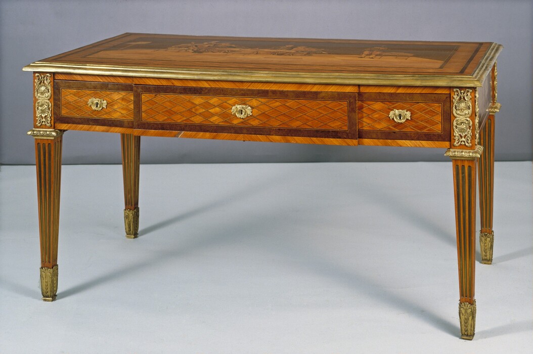 A rectangular golden-brown wood table has three shallow drawers in a row under the top surface, which is inlaid to create a scene, though details are difficult to make out in this photograph. The center drawer is the widest, and all three are inlaid with a horizontal grid of diamonds and outlined with a border of darker wood. Gold-colored metal mounts cap the feet, and gold ornament edges the tabletop and parts of the drawers, including the keyholes and handles. Metal ornaments above the front legs have mirrored palms framed by vines that curve into shapes of harps. The fluted legs taper below the drawers.