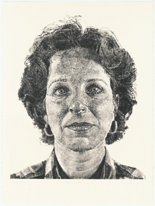 The image is a detailed portrait of the entire head of a person facing directly towards the viewer in a frontal position. The individual has strong facial features, clear eyes, defined lips, short slightly curly hair, and is wearing medium-sized hoop earrings. They are dressed in a collared garment, with a plain background.