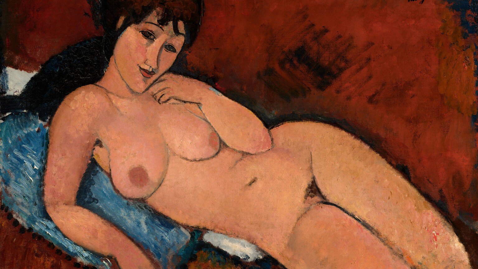 A nude woman with long, dark brown hair and peach-colored skin reclines against a denim-blue cushion in this stylized, horizontal painting. The painting is done mostly with areas of mottled color outlined in delicate black lines. Shown from the knees up, the woman lies with her legs to our right, and her hips tipped forward so her body faces us. She looks at us with dark, almond-shaped eyes under thin, arched brows. Bangs gather across her forehead, and her dark hair seems to be pulled back. Her head is tipped to our right, and she has an upturned nose, smooth cheeks, a pointed chin, and her smiling coral-red lips are closed. She holds the back of her left hand to her cheek and props herself up on her other elbow. Her rounded breasts have rust orange-colored nipples, and the contours of her body are smooth. There is a patch of dark hair at her groin, and her legs are pressed together. The blue pillow she rests on overlaps a white cloth or second pillow. The rest of the background is painted a terracotta brown with loose brushstrokes, so the setting is undefined. The artist signed the painting in dark letters in the upper right corner: “modigliani.”