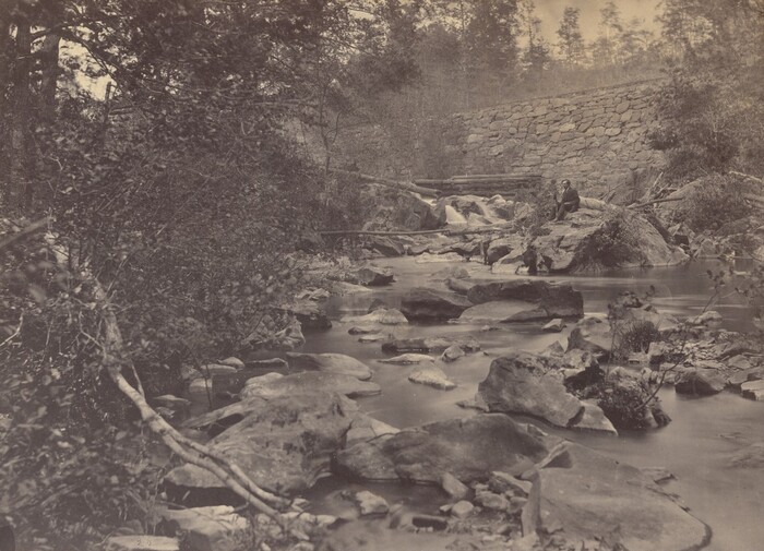 The image portrays a natural landscape with a river flowing over rocks. Dense foliage and a stone wall line the riverbanks. A person sits on a big rock near the water. The photograph uses a long exposure technique, creating a smooth appearance on the flowing water. The color scheme is composed mainly of sepia tones, creating a timeless and serene mood.