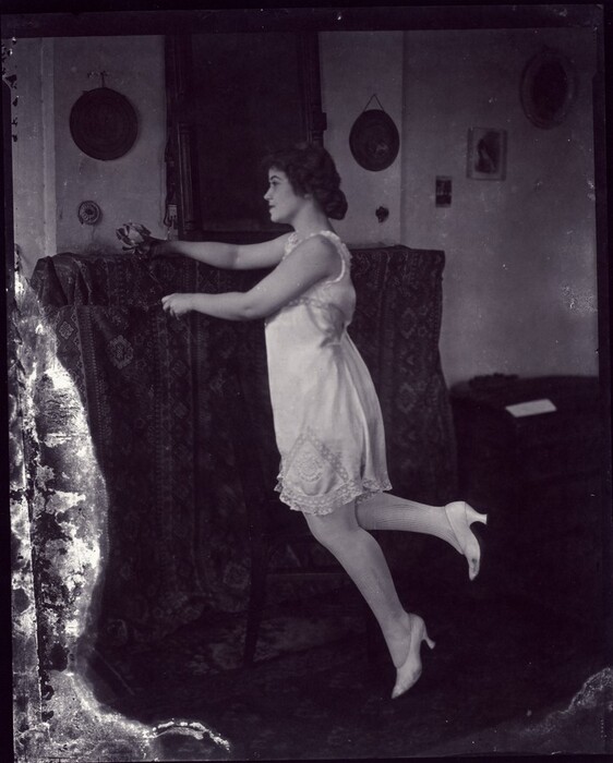 The image shows a woman in profile, standing on one leg while holding onto the back of a chair for balance. She has a facial profile with eyes looking forward, hair styled in a loose updo, and wears a light-colored knee-length sleeveless dress with lace detailing. She also has stockings and pointed shoes with small heels, holding a delicate flower in her hand. The setting appears to be indoors, with patterned fabric draped over the chair and circular decorations on the wall behind her. A cabinet or side table is visible to the right with a small, indistinct object on it. The image shows some wear on the edges.