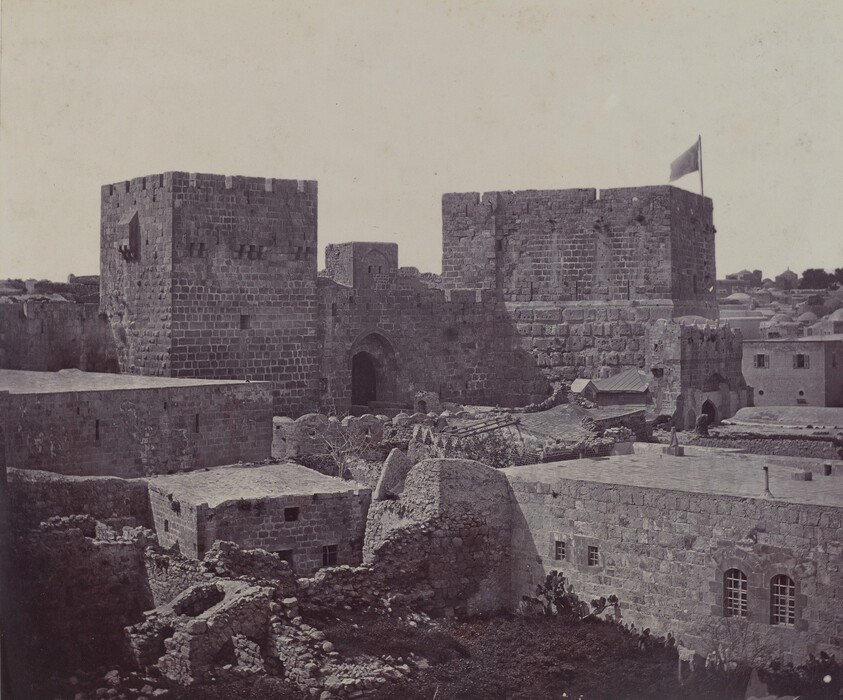 The image shows a historic stone fortification with massive towers and walls built from rectangular stones. The view is from an elevated perspective looking down, showing the structure with a high horizon. A flag waves on the tallest tower on the right. The architecture consists of intact and partial walls, with at least one arched entrance visible. The color palette is monochromatic, varying shades of gray. The image may appear more like a historical photograph with fine details, rather than a painting. Other buildings extend into the distance beyond the stone structures.