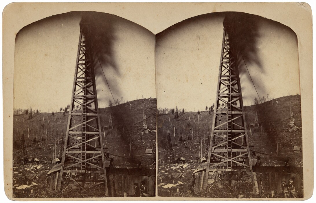 The image shows a stereoscopic view of an oil derrick in a sparse landscape. The point of view is from ground level, looking up at the tall wooden structure. The horizon line is low, with the derrick towering against the sky. In the distance, there are scattered small buildings, additional derricks, and some remaining trees. The overall color palette is a muted sepia tone. Closest to us, the base of the derrick shows wooden structures and two figures. The surrounding area has a hilly terrain with wood debris, showing a human impact on the environment.