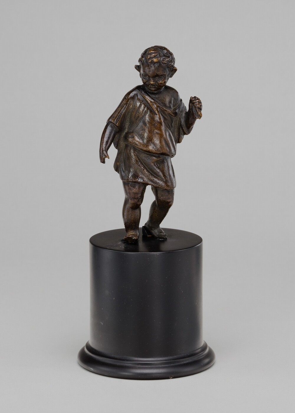 The sculpture depicts a small child standing with a slight tilt. The child is clad in a simple draped garment and has curly hair. The sculpture has a dark, bronze-like appearance with a smooth finish. It stands on a cylindrical black base.