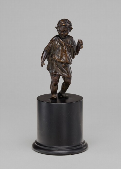 The sculpture depicts a small child standing with a slight tilt. The child is clad in a simple draped garment and has curly hair. The sculpture has a dark, bronze-like appearance with a smooth finish. It stands on a cylindrical black base.
