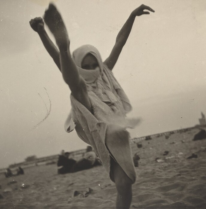 The image shows a person captured from head to mid-thigh, with the person's face mostly obscured by a fabric that covers their head and drapes down. The individual is in a dynamic pose, captured in mid-motion with one leg high in the air as if executing a high kick, and their arms are raised above the head. The person appears to have short hair partially visible underneath the fabric. They are wearing a loose, textured garment that appears to be a towel or similar material, which is notably flowing with their movement. The background is a sandy area, likely a beach, with a few blurred figures sitting or lying on the ground in the distance, under a sky that seems overcast or lightly cloudy.