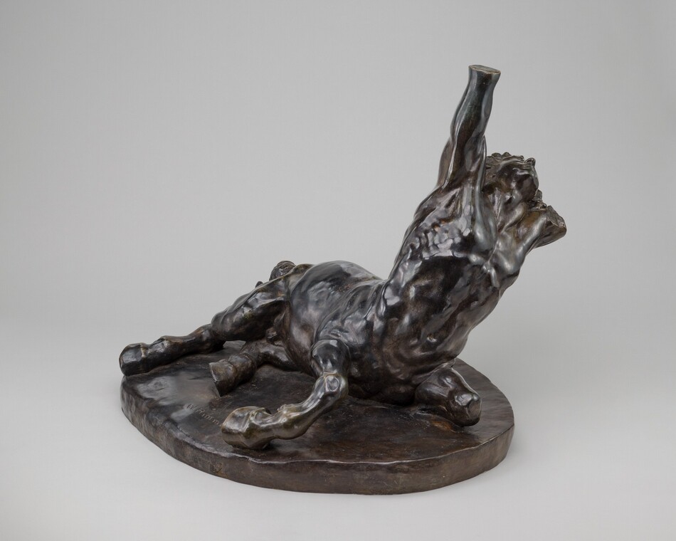 The sculpture shows a figure on its back, legs bent, and an arm raised upwards. The figure appears muscular and is positioned on an irregular base. The sculpture is dark bronze with a metallic sheen. It seems to be made of metal, possibly bronze, giving it a reflective and solid appearance. The sculpture is placed on a flat surface and appears relatively small. The detailed texture of muscles and base is skillfully rendered.