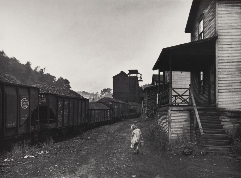 The image shows a dirt path with train cars on the left and a wooden house with a porch on the right. The horizon is unclear and obscured by mist, aligning with the tree tops and train cars. The image is a photograph in black and white, highlighting contrasts in the scene. A child in a light garment is walking on the path near the train cars.