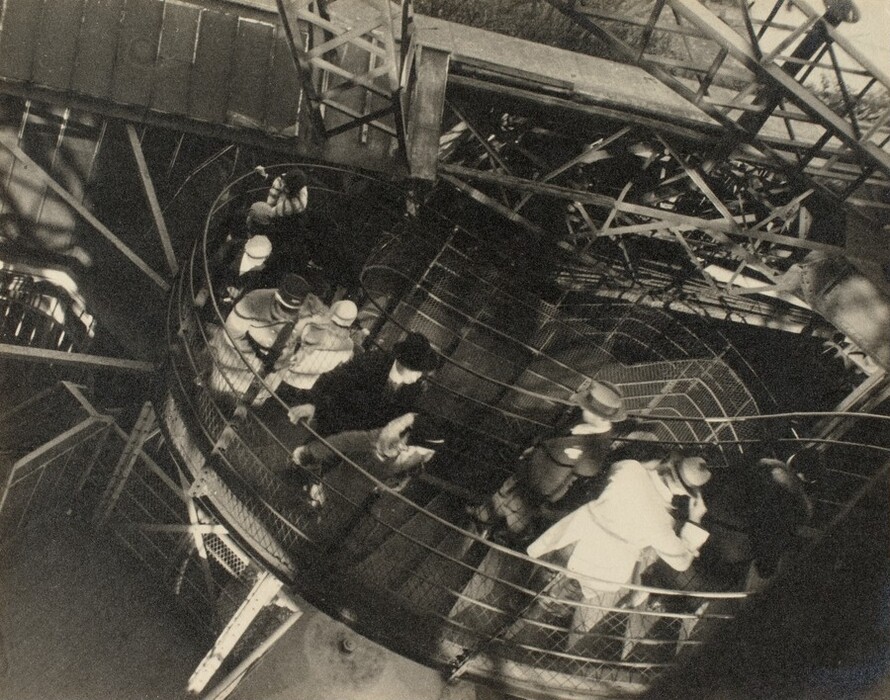 This is a photograph of a group of people walking on a spiral staircase. The image shows an overhead view of a circular, metal mesh-enclosed staircase with intricate metal beams and structures. The staircase spirals downwards. The photograph is in black and white, emphasizing light and shadows. The composition highlights the geometric patterns formed by the stairs and handrails, contrasting with the moving figures.