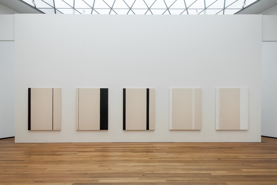 Five abstract paintings hanging on a white wall in a room with hardwood floors and a skylight. 