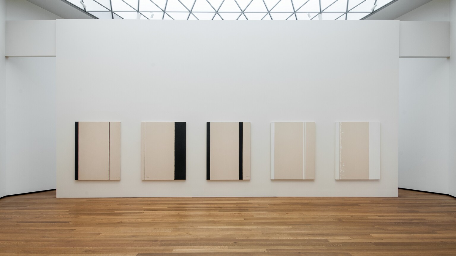 Five abstract paintings hanging on a white wall in a room with hardwood floors and a skylight. 