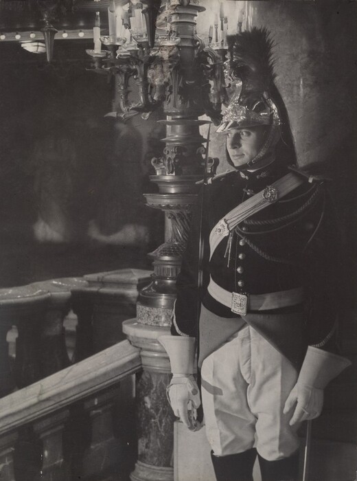 The image shows a man from the knees up, positioned at the corner of a grand staircase. He is standing upright, facing to the side with a serious expression. His facial features are partially obscured by the ornate helmet he is wearing, which is adorned with a prominent plume. The man's hair is not visible due to the helmet. He is dressed in a ceremonial uniform including a dark jacket with decorative buttons, a shoulder sash, and a belt featuring a detailed buckle. His white trousers are tucked into polished boots, and he is wearing a pair of gloves. The man's hands are relaxed by his sides, and he appears to be holding a long object, possibly a ceremonial staff or sword, that rests on the ground. The background reveals an opulent setting with an elaborate candelabra on a pedestal, part of a wide staircase with marble and wooden elements. The lighting suggests a dimly lit interior with reflective surfaces creating subtle highlights.