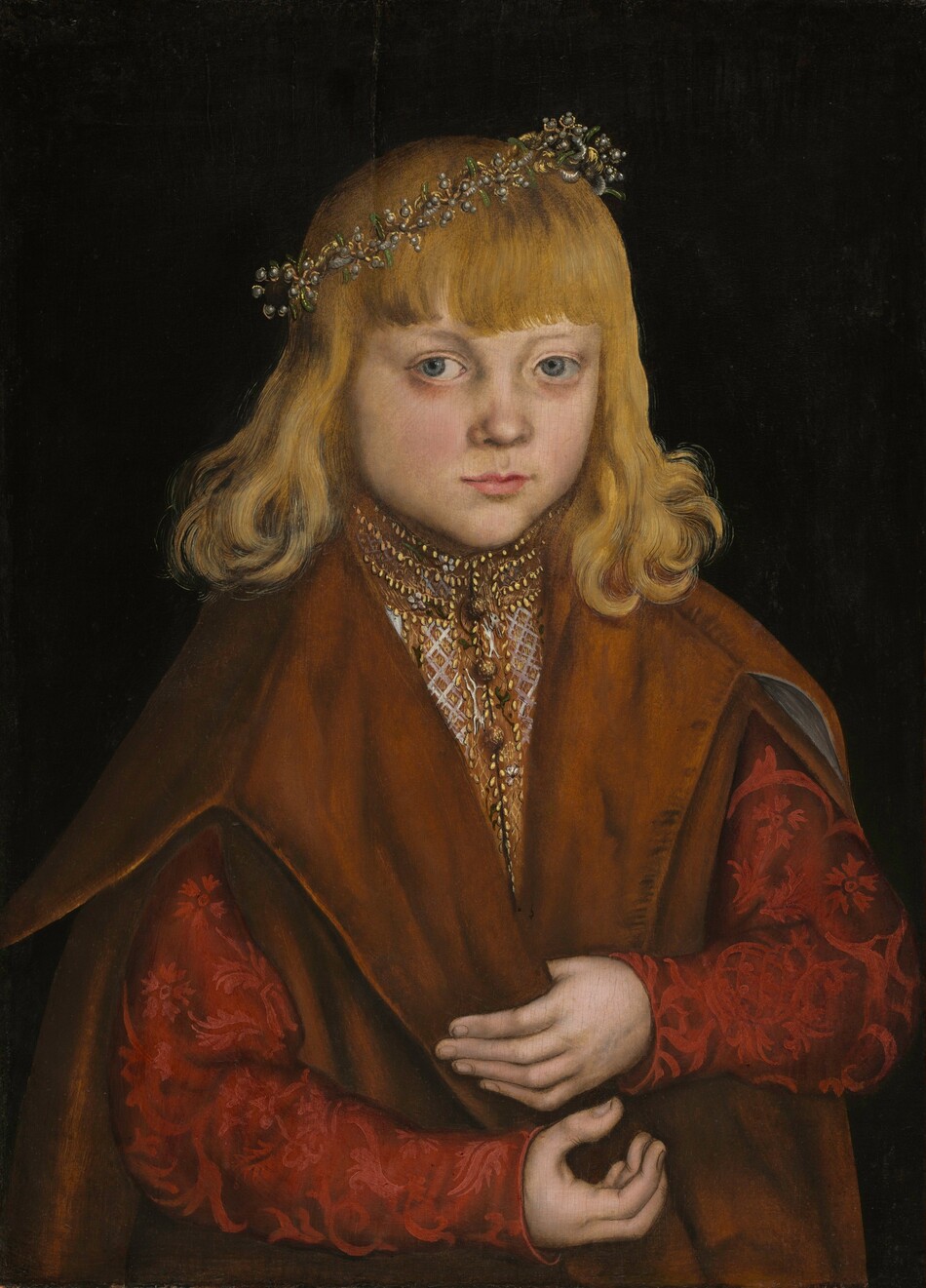 Shown from the waist up, a young boy with blond hair faces us, wearing an embroidered garment and heavy cloak in this vertical portrait painting. His body angles slightly to our right, but he looks just off to our left with pale blue, faintly bulbous eyes. He has pale skin, flushed cheeks, a delicate nose, a pointed chin, and his pink lips are closed. His wavy hair curls around his shoulders, and a fringe of bangs spans his forehead. A crown of black pearls, gold, and green stones is set at an angle on his head. His high-necked light brown shirt is edged with gold and embroidered with white and gold. The long sleeves are brocaded with a ruby-red floral pattern against darker garnet red. He holds the edge of a caramel-brown, sleeveless overcoat with one pale hand, and the other is cupped with fingers pointing upward just below. He is shown against a black background.