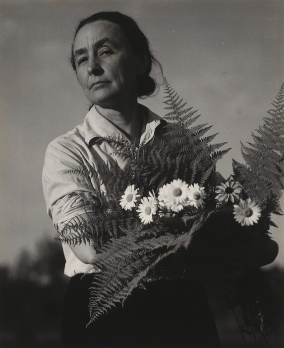 The image depicts a woman standing and facing slightly to the side. She has a contemplative expression with high cheekbones and a furrowed brow. Her dark hair is pulled back tightly. She is wearing a light-colored long-sleeve button-up shirt with partially rolled-up sleeves, and she is holding a bouquet of daisies and ferns. The background shows a cloudy sky and some indistinct foliage or trees in the distance.