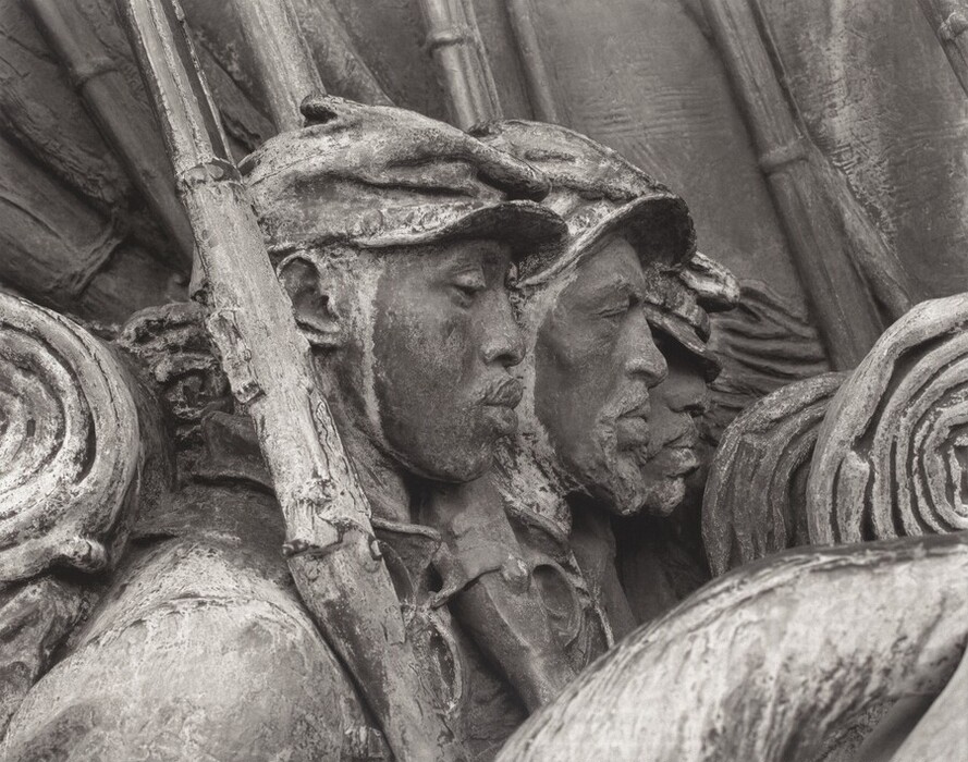 The sculpture depicts a group of soldiers in profile view wearing military attire and carrying rifles. The figures have detailed facial features and the sculpture is monochromatic with a rough texture, suggesting aged metal. The soldiers are closely grouped and the scene conveys a sense of movement and unity.