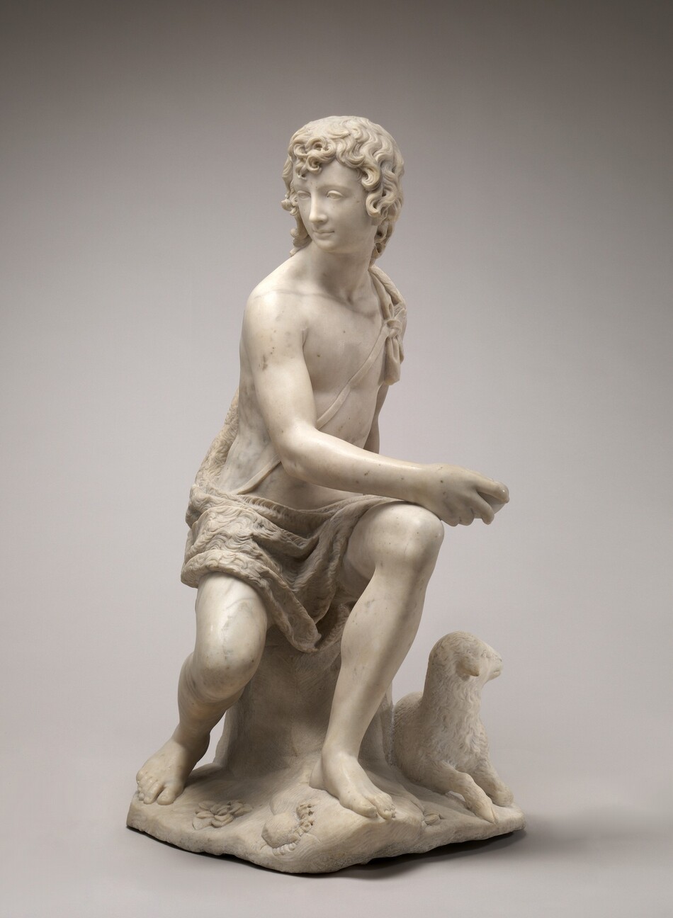 This sculpture depicts a young male figure in a seated pose, with his body facing right and his head turned over his shoulder towards the left. He is wearing a draped garment that covers one shoulder and goes behind his back, leaving the other shoulder bare. The fabric pools in his lap and is tied by a string across his chest. The young man has curly hair and a straight nose, and he wears a neutral expression. He appears to hold a stone or round object in one hand, which he rests on the opposite knee. He seems to sit on a rock or tree stump, and beside him on the right is a small lamb that looks off to the right. Below his feet is a small area of ground with some small rocks and flowers. The sculpture is made out of a white material with some gray marbling.