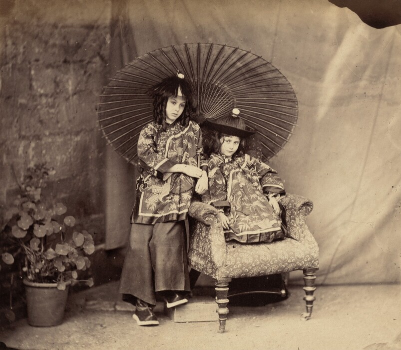Two young girls with shoulder-length, dark hair wear Chinese, mandarin-style tunics in this nearly square black and white photograph. Both appear to have pale skin. They have short, rounded noses and dark eyes, and their lips are closed. One girl sits with her legs tucked under her on an upholstered armchair as she looks steadily out at us. The other, to our left, stands and leans onto one elbow, which is propped on the back of the chair. She reaches her other arm across her body to rest her hands near each other. Her hair falls in long, defined curls. She also wears a mandarin-style tunic and wide-legged pants. She rests one foot up on a block partially tucked under the chair, so her knees are together. She looks down and off to our right. Both tunics are decorated with long-necked birds and plants. The girls wear conical-shaped hats, each with a small white ball at the top. A wide parasol with closely spaced ribs is open behind the pair. The space behind them appears to be draped with cloth, and a plant in a pot sits to the left.
