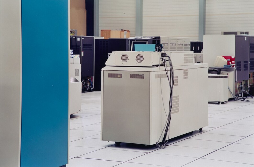 This is a photograph of computer equipment in a server room. The image shows a room filled with large box-like pieces of computer equipment and various consoles. There is a main console in the foreground, with a light gray exterior and multiple cables connected to it. The floor is tiled, typical of data centers. In the background, additional computing units are arranged along the sides of the room, with some having protruding wires and elements. The space is well-organized, appearing functional and sterile, suitable for technology management.