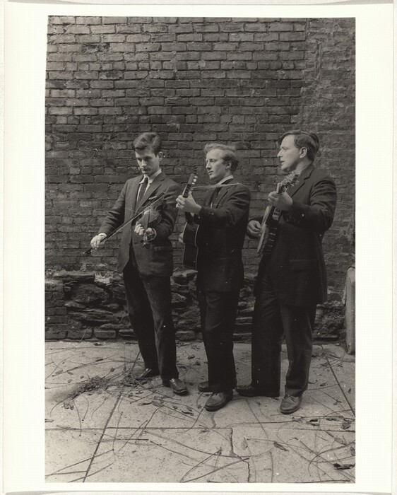 There are three men standing against a brick wall. The man on the left is playing a violin, the man in the middle is playing an acoustic guitar, and the man on the right is playing a banjo.