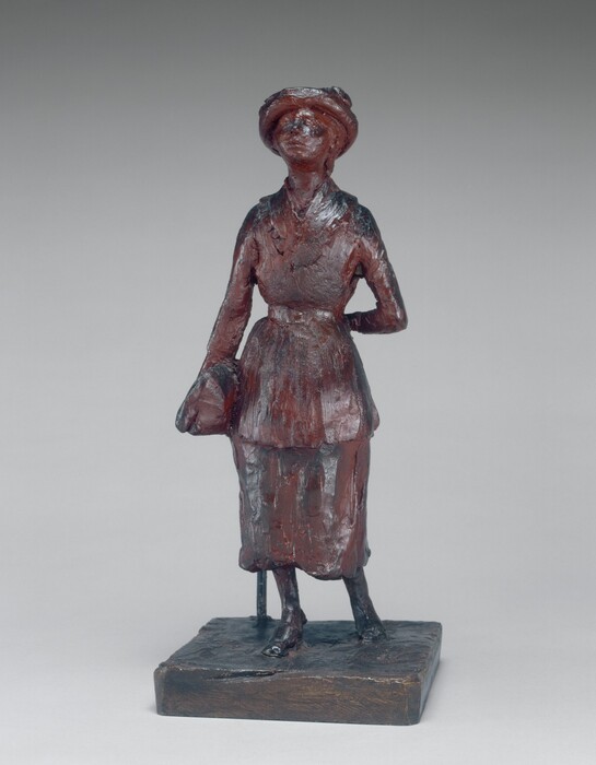 The sculpture depicts a standing figure of a woman dressed in early 20th-century attire. The woman is wearing a long skirt and a blouse with a belt at the waist, and she has a wide-brimmed hat on her head. The sculpture is dark reddish-brown and appears to be made of a material resembling bronze, with a textured surface capturing the details of the clothing and posture. The woman is standing upright with one hand behind her back and the other holding onto a small handbag. She is mounted on a simple rectangular base, and the overall sculpture is proportional to a small human figure, suggesting a scale model of an everyday person of that era.