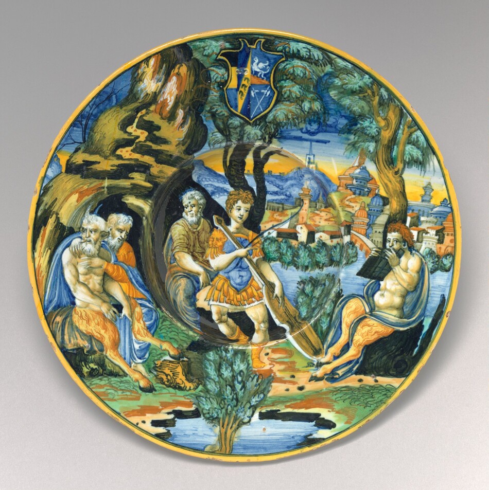 This is a painting of a decorative ceramic plate featuring a mythological scene. The plate is decorated with classical figures in an outdoor setting. The central figure is a person in classical attire with a blue and orange outfit, holding a spear or staff. To the left are older, bearded figures seated under a large tree. On the right, another figure sits under a tree, possibly reading from a scroll. The background shows a landscape with a walled city, turrets, and a river winding through greenery. The upper part of the plate features a heraldic shield with a yellow border. The style reflects the Italian Renaissance era.