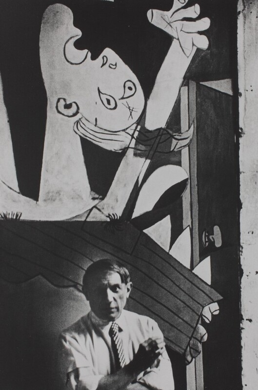 A man with a round face and straight, ear-length hair stands at the foot of a mural that fills the background of this vertical black and white photograph. The man looks at us with shaded eyes, and he wears a light-colored collared shirt with the sleeves rolled up to the elbows. He wears a checked tie and crosses his arms, holding one cupped hand up as if shielding a cigarette from a breeze. The abstracted mural behind him shows a person with their face tipped up and one hand raised. The mouth and eyes are wide open. The person’s features are painted with dark outlines against a dark background.
