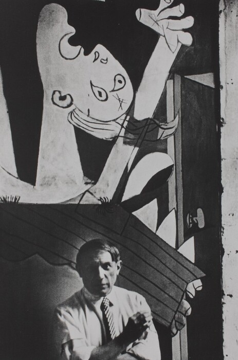 A man with a round face and straight, ear-length hair stands at the foot of a mural that fills the background of this vertical black and white photograph. The man looks at us with shaded eyes, and he wears a light-colored collared shirt with the sleeves rolled up to the elbows. He wears a checked tie and crosses his arms, holding one cupped hand up as if shielding a cigarette from a breeze. The abstracted mural behind him shows a person with their face tipped up and one hand raised. The mouth and eyes are wide open. The person’s features are painted with dark outlines against a dark background.
