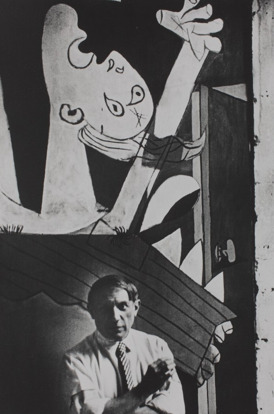 A man with a round face and straight, ear-length hair stands at the foot of a mural that fills the background of this vertical black and white photograph. The man looks at us with shaded eyes, and he wears a light-colored collared shirt with the sleeves rolled up to the elbows. He wears a checked tie and crosses his arms, holding one cupped hand up as if shielding a cigarette from a breeze. The abstracted mural behind him shows a person with their face tipped up and one hand raised. The mouth and eyes are wide open. The person’s features are painted with dark outlines against a dark background.