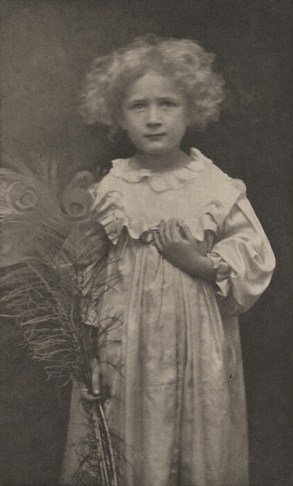 The image shows a young child captured from the waist up in a standing position. The child has curly light hair that frames their face. Their facial features appear soft and calm, with eyes looking directly at the camera. The child is wearing a loose-fitting gown with a ruffled collar and long sleeves. They hold a bundle of peacock feathers in their hands, which are positioned around the waist area. The backdrop is a dark, indistinct background.