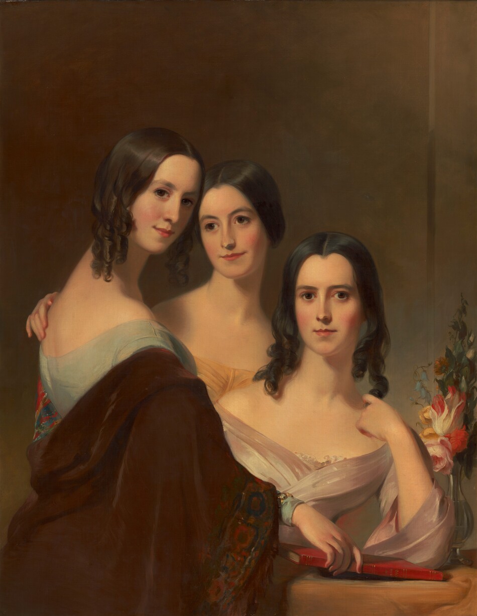 In this painting, three women are depicted standing and sitting closely together. The women have fair complexions and dark hair in ringlets, and they are smiling slightly. The woman on the left appears to be standing, and her body is turned to the right while her head is turned over her shoulder to face us. She wears a light blue dress with a brown shawl, and she is holding the edge of a red book that rests on a small table in the bottom right corner. The woman in the middle also appears to be standing, and she is wearing a golden-yellow off-the-shoulder garment. One of her hands is on the shoulder of the woman on the left. The woman on the right is seated at the small table, and she wears a pale pink off-the-shoulder dress. She looks directly out at us. The small table is covered in a golden cloth, and on the right is a vase of colorful flowers. The wall behind the woman is a blend of brown, tan, and pale blue tones.