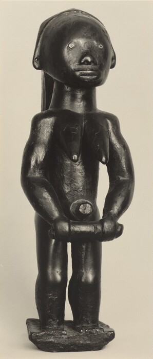 The sculpture depicts a human figure standing upright. The figure has distinct facial features with circular eyes and a prominent nose. The body has broad shoulders and a noticeable chest, with arms bent at the elbows holding an object. The sculpture is dark in color and has a glossy finish, and it stands on a rectangular base. The figure has a solid and simplified form with a slightly uneven texture.