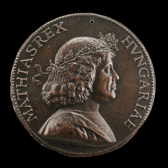 The sculpture is a profile portrait of Matthias Corvinus, the King of Hungary, with the inscription "MATHIAS REX HVNGARIAE" around the edge of the medallion. It is made from bronze, with a dark brown, metallic appearance and a slightly weathered texture. The figure has curly hair with a laurel wreath, and detailed carvings of the face, hair, and clothing folds. Matthias Corvinus faces to the right in a historical and regal portrayal. The medallion resembles a coin or commemorative medal.
