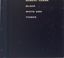 Lettering on this black square book has four lines of white text with “ROBERT FRANK” across and slightly cut off by the top edge. The next three lines together read “BLACK AND WHITE THINGS.” The left edge of the text is lined up left of center of the cover, and there are three holes punched on the leftmost edge, suggesting it might be part of a binder or notebook. The cover has some scuffs and dings.