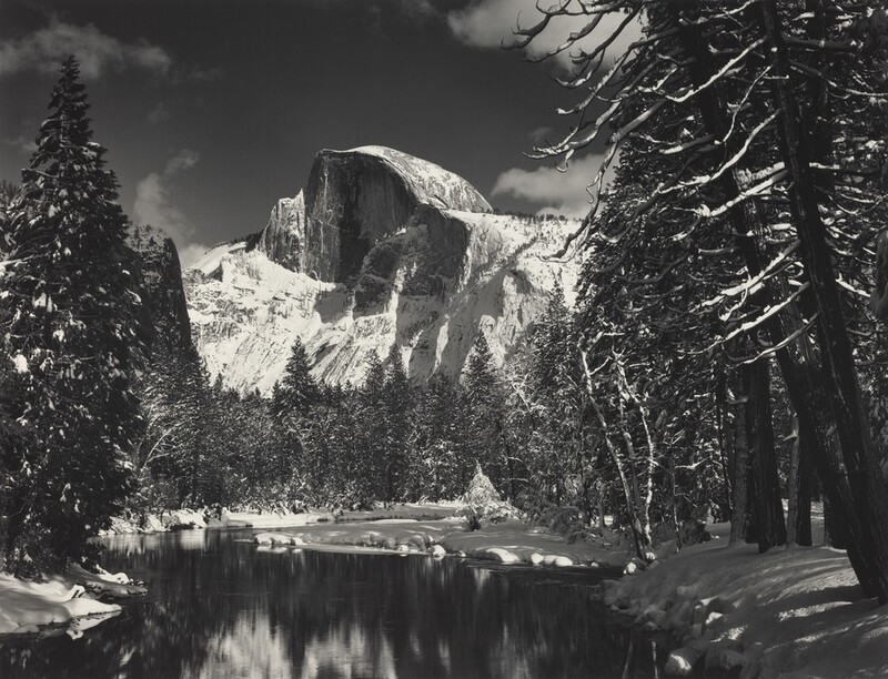 Across a still, reflective lake, a curving rock formation juts high into the sky in this horizontal, black and white photograph. Snow-dusted trees, both evergreen and the leafless branches of deciduous trees, frame the scene and line the far side of the lake. Snow blankets the rocky ridges around the Half Dome, the flat face of which is angled to our left. A few puffy clouds drift across the sky.