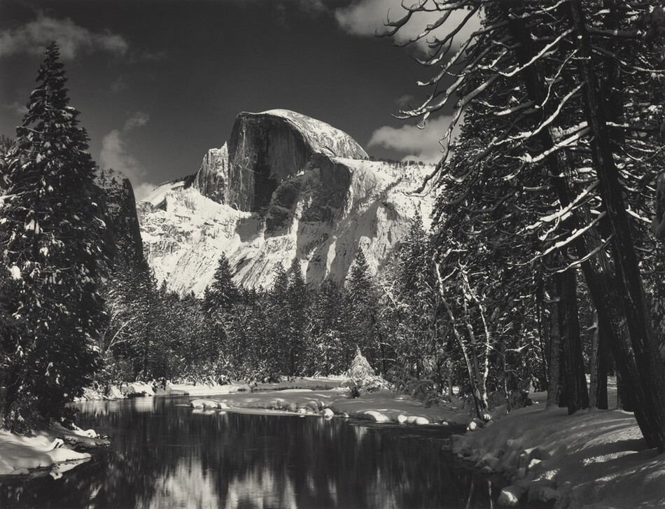 Across a still, reflective lake, a curving rock formation juts high into the sky in this horizontal, black and white photograph. Snow-dusted trees, both evergreen and the leafless branches of deciduous trees, frame the scene and line the far side of the lake. Snow blankets the rocky ridges around the Half Dome, the flat face of which is angled to our left. A few puffy clouds drift across the sky.