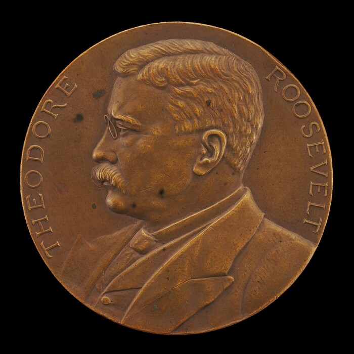 The image shows a round medallion with a profile relief of a man with a mustache and glasses, dressed formally. The man depicted is Theodore Roosevelt, as indicated by the engraved text "THEODORE ROOSEVELT" along the edges of the medallion. The sculpture is cast in a bronze-colored material with variations in tonality. The detail of the sculpture reflects a realistic representation, with features like combed hair and suit folds. The medallion emphasizes Roosevelt’s profile.